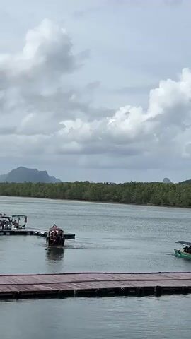 A long-tail boat with multiple passengers departs from a pier on the left side of the frame, moving away from the camera and towards the right. Another smaller boat is visible in the distance to the right. The pier itself is composed of dark, flat planks. The water appears calm and reflects the cloudy sky. In the background, a dense green forest lines the shore, with rolling hills or mountains visible beyond the treeline. The sky is overcast with dramatic clouds.