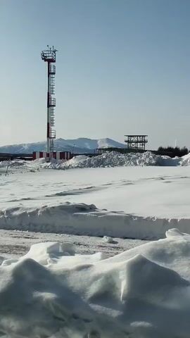 The scene is a snow-covered landscape under a clear, bright sky. A tall, red and white striped tower stands prominently in the middle ground, next to a small building with the same red and white stripe pattern on its roof. In the background, there are snow-capped mountains and a metal structure. The foreground is filled with snow, with visible tire tracks on the ground. The camera seems to be moving slightly, as if from a moving vehicle.