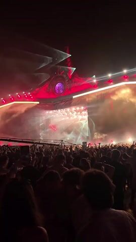 A crowded outdoor concert at night. A massive stage structure dominates the background, illuminated with red and white lights and smoke effects. A large screen displays abstract visuals. In the foreground, a dense crowd of people are visible, many with their hands raised, enjoying the performance.