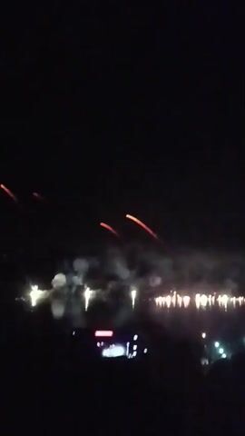 The video shows a nighttime scene of a fireworks display over a body of water. In the foreground, silhouettes of people are visible, with one person holding up a phone to record the event. The fireworks are exploding in the distance, creating bursts of light and smoke that are reflected in the water. Several bright lights, possibly from boats or other structures, are also visible along the shoreline. The overall atmosphere suggests a celebratory event.