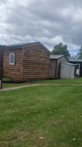 A concrete path leads through a grassy area where several small, prefabricated structures are located. The structures appear to be part of a campground or mobile home park. On the left, a small wooden building with a metal roof and a single window is visible. Next to it is a corrugated metal structure with a red fire alarm box mounted on the side and a dark brown door with a number '2' on it. Further down the path, more of these structures are visible, including what appears to be a mobile home with a ribbed exterior. The sky is partly cloudy.