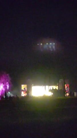 The video shows a concert stage at night. There are bright lights illuminating the stage and screens, as well as a crowd of people watching the performance. The sky above the stage is dark, with several spotlights visible. There are also some trees to the left of the stage that are lit up with purple lights. The overall atmosphere is that of an outdoor music festival.
