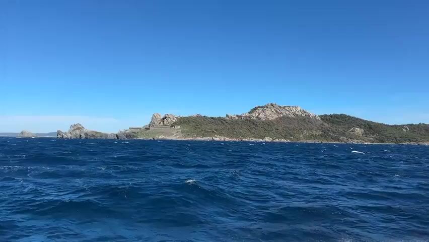 The video shows a vast expanse of deep blue, choppy water under a clear blue sky. In the distance, a rocky, green-covered island juts out from the sea. The island features rugged, light-colored rock formations and appears to be largely uninhabited, with some structures visible, possibly ancient ruins or fortifications, scattered along its rocky coastline. The perspective suggests the video was filmed from a moving boat, as the waves are clearly visible and appear to be a constant feature. The overall scene evokes a sense of isolation and natural beauty.