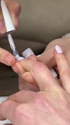 A person is applying light purple nail polish to the fingernails of another person's left hand. The manicurist is holding the person's hand with their left hand while using their right hand to paint the nails. The manicurist's hand is slightly red and shows visible knuckles. The nail polish is a pale lavender color and comes in a small white bottle. The background is slightly blurred, suggesting a close-up shot.