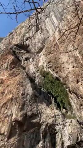The video shows a white, multi-story building by a river, nestled against a large rock cliff face. The cliff is tall, rough, and textured, with some greenery growing on it. The river is a clear, blue-green color. The building has multiple windows and a dark roof, with a stone wall bordering the river. The sky is bright and blue. The camera pans slightly from the cliff to the building and back again.