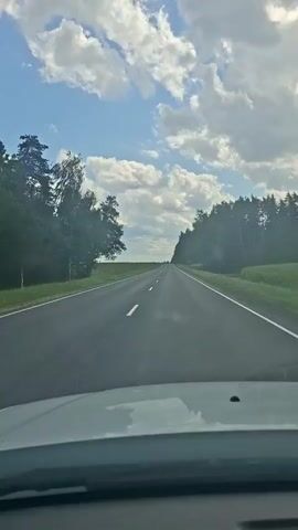 The video shows a view from inside a moving vehicle. The road ahead is asphalt with white dashed lines indicating lanes. On either side of the road, there are fields and trees. The sky is blue with scattered white clouds. The car appears to be traveling on a rural highway.