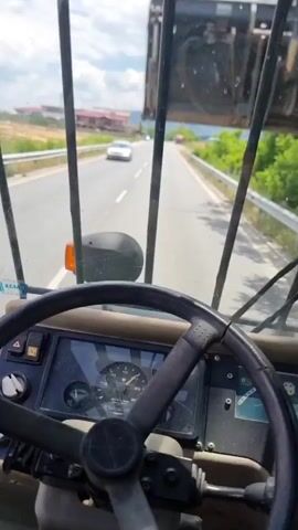 The video shows a point-of-view perspective from inside a moving vehicle, likely a large truck or heavy machinery, driving on a road. The dashboard and steering wheel are visible in the foreground, and the road ahead is visible through the windshield. Above the windshield, a large, dark, metallic structure, possibly part of a crane or industrial equipment, is seen moving. The vehicle is on a paved road with a white dashed line in the center. A white car is visible on the opposite side of the road in the distance. Lush green foliage and trees line the right side of the road, and a metal guardrail runs alongside it. On the left side of the road, buildings are visible in the distance under a blue sky with some clouds. The camera shakes slightly, indicating movement.