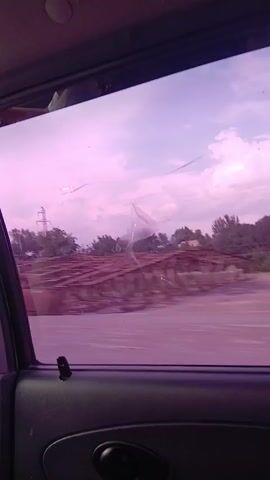 The video captures a view from inside a vehicle looking out the passenger window. The window is tinted purple and appears to have some damage or obstructions, possibly cracks or debris. The car is in motion, indicated by the blurred landscape outside. The scene outside includes a road, trees, power lines, and some buildings, suggesting a rural or semi-urban environment. Several large, white, cylindrical objects are visible along the side of the road, possibly bales of hay or similar agricultural materials. The sky is overcast with a pinkish hue, possibly due to the lighting conditions or the window tint. The interior of the car is visible at the bottom of the frame, showing the door panel and window controls.