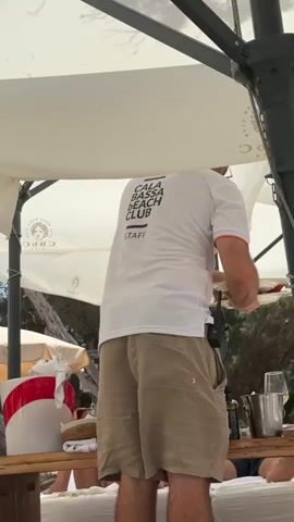 The video shows a beach club environment under large white umbrellas. A person wearing a white t-shirt with "CALA BASSA BEACH CLUB STAFF" printed on the back and khaki shorts is standing near a wooden table or bar. They are facing away from the camera and appear to be holding something in their hands, possibly preparing a drink or serving a customer. On the table, there are ice buckets, bottles, and other items associated with a bar service. In the background, several people are visible sitting or lounging on what appear to be beach beds. The overall scene suggests a busy day at a beach resort, with staff attending to guests. The environment is sandy, with trees and foliage visible behind the beach club setup.