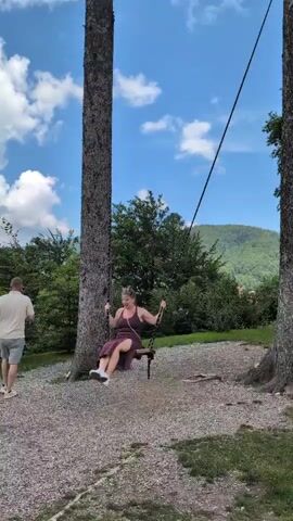 A woman is swinging on a swing suspended between two large trees. She is wearing a dark-colored dress and white shoes. Her hair is pulled back, and she is wearing glasses. A man in a light-colored shirt and shorts is walking behind her, to the left of the frame. The swing is made of dark material and appears to be suspended by ropes or chains. The ground beneath the swing is covered in small rocks and gravel. In the background, there are more trees and a green hillside under a blue sky with white clouds. The lighting suggests it is daytime.