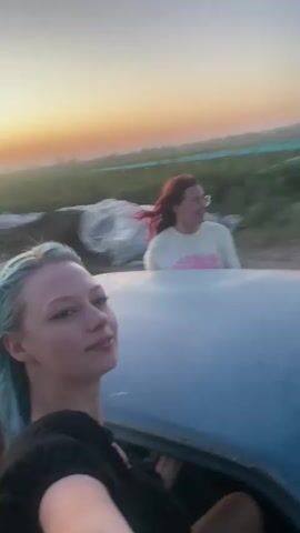 This video shows two young women in a light blue car, possibly a convertible, driving through a rural landscape at sunset. The woman in the foreground, with light blue hair, is leaning out of the car window and looking to her left. She is wearing a black top. Behind her, another woman with red hair, wearing a light-colored sweatshirt with pink detailing, is visible inside the car, looking towards the right. The background features green fields, trees, and rolling hills under a pale orange and blue sky. The car appears to be in motion, as indicated by the blurred background and the dynamic framing of the shots.