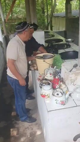 Two men are standing in an outdoor kitchen area. The man on the left is wearing a light-colored polo shirt, blue jeans, and a traditional cap. He is looking at a yellow bucket with a metal handle that is being held by the man on the right. The man on the right is wearing a dark shirt and a traditional cap. He is holding the bucket and appearing to be pouring something from it. In front of them on a tiled surface is a collection of teapots, some plain white and others decorated with floral patterns. Behind the men, there are large flat cooking surfaces that appear to be built-in ovens or stoves. The environment is rustic, with trees and foliage visible in the background.