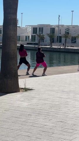 Two people are dancing on a wooden dock in front of a body of water, while a person in a red shirt films them. The dancers are in activewear; one is wearing a pink long-sleeved shirt and black shorts, while the other is in a black top and pink shorts. A tripod stands next to the person filming. Palm trees and a white building with a modern design are visible in the background. The ground is paved with grey tiles.