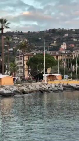 The video shows a waterfront view of a town in Italy.  The camera is positioned on the water, looking towards the shoreline. In the foreground, there are rocks and calm water with gentle ripples.  A low yellow bridge or walkway is visible along the coast.  The town is characterized by closely packed, colorful buildings, predominantly in shades of yellow, orange, and pink, with architectural styles that suggest an older European setting.  Palm trees and other greenery are interspersed among the buildings.  The town extends up a hillside, where more houses and vegetation are visible.  The sky is overcast with a mix of white and grey clouds, creating a soft, diffused light over the scene.  The overall impression is of a quiet, picturesque coastal town at dusk or dawn.