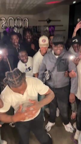 A group of approximately 15 young adults are gathered in what appears to be an indoor party. In the foreground, a man with braided hair, wearing a short-sleeved white shirt and dark pants, is actively dancing. His movements are energetic, and he is the center of attention. The other individuals are standing around him, some holding up their phones, presumably recording or taking pictures of the dancer. Most of the attendees are Black. The room is decorated with silver balloon numbers "2000" visible in the background. There is a patterned ceiling with recessed lighting. The overall atmosphere is lively and celebratory.