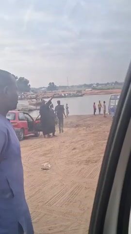 The video shows a sandy area beside a river. There are several people, a red car, and boats on the river. The people are mostly gathered near the riverbank. Some people dressed in dark clothing are near the red car. They seem to be assisting someone in dark robes to walk. The red car has its driver-side door open. There are tire tracks in the sand, and the sky is overcast. The video is taken from inside another vehicle.