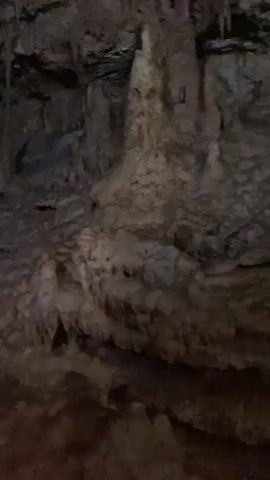 The camera pans across a cave with limestone formations. The cave has large stalactites and stalagmites. A group of people can be seen walking on a path through the cave. The cave is dark and lit by artificial lighting.