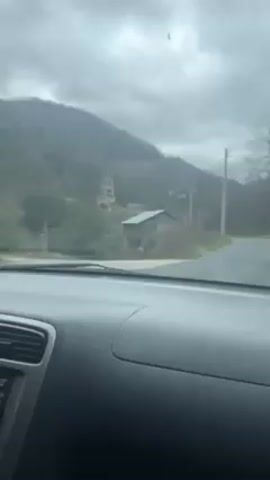 The video shows a view from inside a car as it drives down a rural road on a cloudy day. In the distance, a mining structure is visible on a hillside. The road has power lines running alongside it. There are some small buildings to the side of the road. A dark-colored SUV is parked near the mining structure. The dashboard of the car is visible in the foreground.