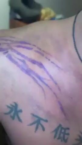 The video shows a shirtless man with a purple tattoo design sketched on his left shoulder and chest. He also has a tattoo of four Chinese characters on his chest. He is wearing a necklace with a pendant. In the background, there's a dresser with various objects, including a water bottle and clothing. The man moves slightly, showing more of the tattoo design and his chest.