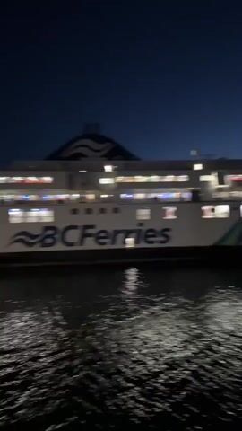 The video, taken at night, shows a BC Ferries vessel docked at a terminal. The water reflects the lights from the ferry and the terminal, creating shimmering patterns on the surface. The camera appears to be on another vessel, providing a view of the docked ferry from its side. Trees and lights can be seen on the shore in the background. The overall atmosphere is dark, with the lights providing the only significant illumination.