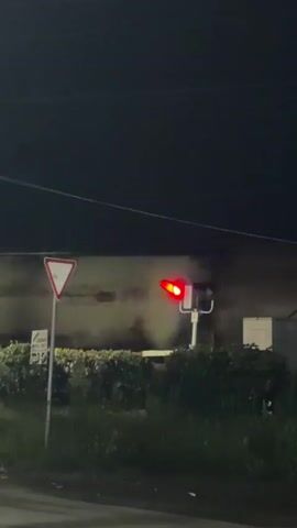 The video captures a dark, outdoor scene at night, likely near a road or railway crossing. A traffic light for vehicles is visible in the center of the frame. It cycles through its lights, first off, then showing two red lights, and then off again. To the left of the traffic light, a white triangular yield sign with a red border is mounted on a pole. Behind the yield sign is a building or structure, its details obscured by darkness. A dense hedge or shrubbery runs horizontally across the middle ground, separating the road from the area with the traffic light and structure. The ground is covered in grass and other vegetation. In the background, the sky is completely dark, indicating nighttime. The overall impression is of a quiet, unattended area at night, with the traffic light functioning as a signaling device.