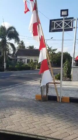 The video shows a red and white flag flying from a makeshift bamboo pole on a sunny day. The flag is partially obscured by the pole and appears to be made of two horizontal stripes, red on top and white on the bottom. The flag is torn and frayed in some areas, suggesting it has been exposed to the elements for some time. The background of the video shows a paved road, a house with a white roof, and palm trees. A motorcycle is visible in the distance, and a street light is mounted on a pole behind the flag. The overall scene appears to be a rural or suburban area.