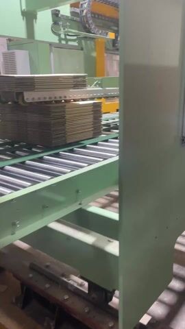 The video shows an automated process involving stacks of cardboard. The stacks are on a roller conveyor, moving past a piece of machinery enclosed by transparent panels. Hoses, wires, and other parts are visible within the machine. The machine appears to be lifting, manipulating, or otherwise processing the cardboard stacks. The machine's control panel is visible through the glass. The machine and the conveyor are both painted a light green color. The floor is concrete, and light is reflecting off the conveyor rollers.
