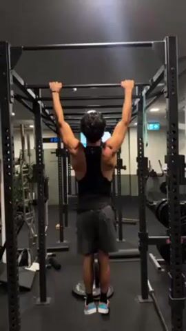 A man is performing pull-ups in a gym, filmed from behind. He is wearing a black tank top, dark shorts, and a weightlifting belt around his waist. His dark, curly hair is visible. He is gripping a pull-up bar with an overhand, wide grip, and his body is hanging straight down with arms fully extended at the beginning of the exercise.

As he performs the pull-ups, his back muscles, particularly his lats and deltoids, are clearly visible and well-defined, contracting as he pulls himself upwards. He executes two full repetitions. In each repetition, he pulls his body up until his chin is well above the bar and his upper chest approaches the bar, demonstrating good range of motion. He then lowers himself back down in a controlled manner, fully extending his arms at the bottom of each rep.

The gym environment features black metal power racks or rigs. Bright overhead lights illuminate the area. In the background, other gym equipment can be seen, along with a green "EXIT" sign on a wall. A weight plate is visible on the floor directly beneath where the man is hanging, though he is not using it for weighted pull-ups in this clip. Background music is audible throughout the video. The man's form appears controlled and strong.