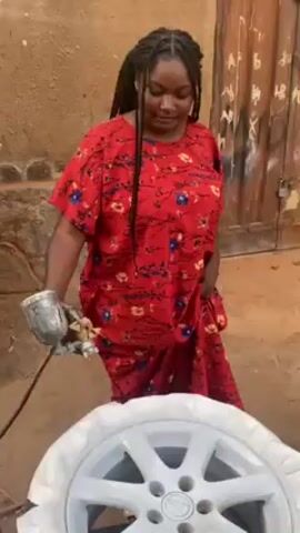 A woman in a red patterned dress with her hair in braids stands outdoors painting a car wheel white using a spray gun. The wheel is sitting on a tire. A weathered wall and a closed door with the word “XABA” written on it form the backdrop. The woman appears to pause and wipes her forehead before resuming the painting.