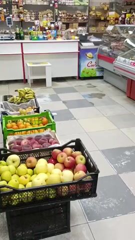The video shows the interior of a small grocery store. In the foreground, there are several crates of fruit, including apples, peaches, oranges, and bananas. Behind the fruit, there are refrigerated display cases filled with various food items such as dairy products, meats, and desserts. Further back, shelves are stocked with packaged goods, including cookies and drinks. The store appears to be well-lit, and the overall impression is one of a typical neighborhood grocery store.