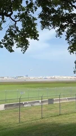 A plane is shown ascending into the sky above an airport tarmac. The plane is white with a red tail. Several other planes and airport buildings are visible in the background. The foreground shows a grassy field with a chain-link fence. The sky is partly cloudy and blue.