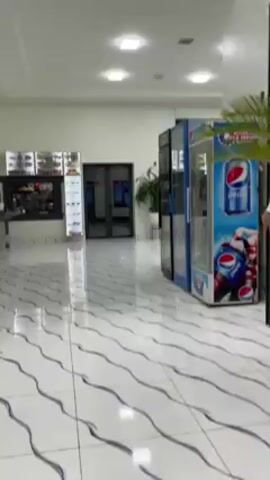 The video shows the interior of a brightly lit commercial space with tiled floors and white walls. The camera pans from a hallway with vending machines to a food service area labeled "Keddhuszel!". The food service area contains stainless steel counters and food displays. People are visible in the background.