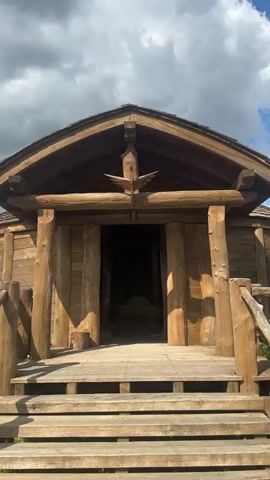 The video shows the entrance to a wooden structure that appears to be a historical dwelling or exhibit. The structure is made of rough-hewn logs and has a thatched roof. There are wooden steps leading up to the entrance, and a wooden doorway. Inside the structure, there are several wooden posts supporting the roof, and a rough stone or concrete floor. In the center of the room, there is a raised platform with what appears to be a throne or a seat. Various objects are scattered on the floor, including what look like animal hides or rugs. The overall impression is of a rustic and ancient setting.