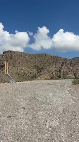 The video shows a roadside scene in a mountainous area under a blue sky with scattered white clouds. In the foreground, there is a gravelly, uneven surface. To the left, a yellow structure with a ramp or stairs leads upwards. Further back, a road curves around a mountainside. A red and white chevron sign indicates a sharp turn to the right. A silver car drives around the curve on the road. Another silver car is partially visible on the right side of the road. The mountains in the background are rocky and covered with sparse vegetation.