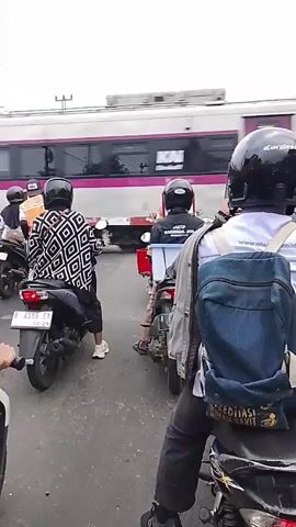 Several motorcyclists wearing helmets wait at a railway crossing as a train passes. The motorcyclists are facing away from the camera, and the train is moving from left to right. The train is white with pink and blue accents. The motorcyclists are on a paved road, and the sky is overcast. One motorcyclist in the foreground is wearing a blue backpack.
