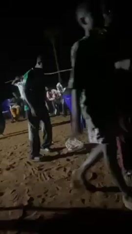 The video, taken at night, shows a gathering of people on a sandy ground. Several individuals stand around, some are seated in blue plastic chairs.  There's a string strung between posts, possibly forming a makeshift barrier.  One person wearing a green shirt and dark pants is dancing.  The lighting is dim, making it hard to discern specific details.  The scene appears to be an informal outdoor event.