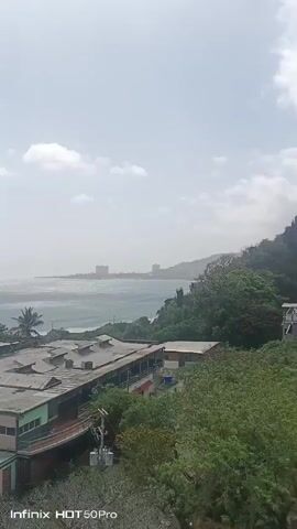 This media file presents an aerial view of a coastal landscape in El Tigrillo, Venezuela. The scene captures a body of water, likely the ocean, stretching towards the horizon under a bright, hazy sky. The water exhibits varying shades of blue and turquoise, with gentle waves breaking near the shore.

Dominating the foreground are several buildings with flat roofs, appearing to be part of an institutional or educational complex. These structures feature a combination of brick and stucco exteriors, with some showing signs of wear. Greenery, including trees and what appear to be palm trees, surrounds the buildings and drapes over the hillsides. A paved road with yellow and white markings winds along the coastline in the middle ground, with a dark-colored vehicle visible on it.

The time of day appears to be daytime, likely late morning or afternoon, given the brightness of the sky. The weather seems warm and possibly humid, as indicated by the haze that softens the distant view of the coastline and any structures further inland. The overall atmosphere is tranquil, suggesting a peaceful, possibly tropical, environment.

Visible text at the bottom of the frames reads "Infinix HOT 50Pro," indicating the device used to capture the media. There are no discernible people in the footage. The activity is passive, focusing on the scenic view of the coast and the surrounding environment.