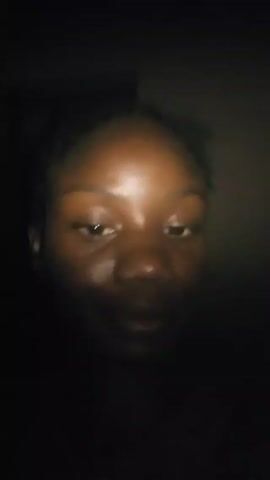 A person with dark skin is centered in a very dark environment. The person looks directly at the camera and smiles slightly. The person's face is illuminated enough to see details, including eyes, nose, and mouth. The person appears to be wearing a dark-colored shirt. The background is completely black.