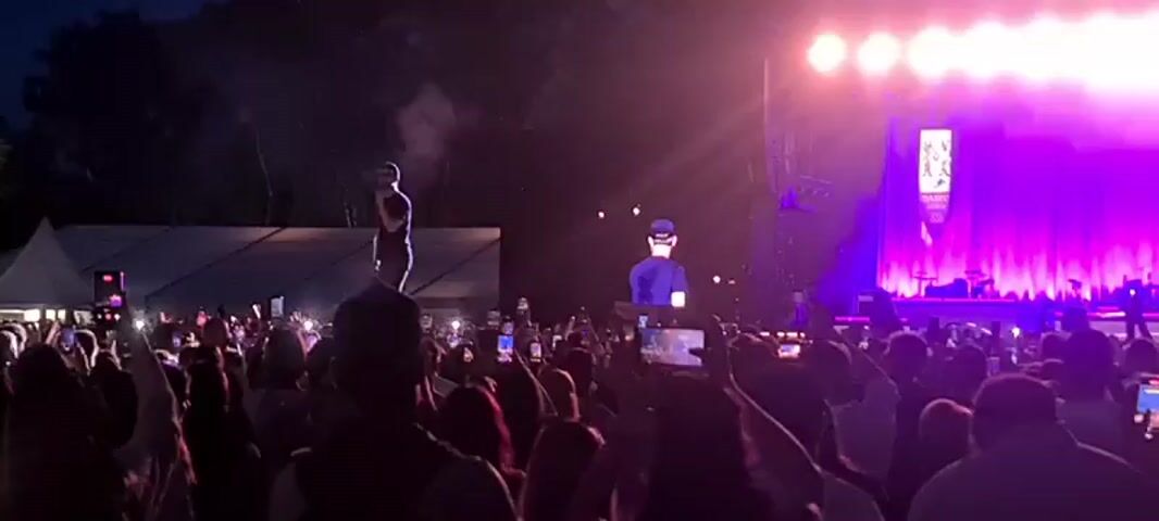 The video shows a concert at night with a large crowd of people in the foreground. A performer is on a stage, standing on a platform and holding a microphone. Behind the performer, a large screen displays a close-up of hands playing a keyboard. The stage is lit with purple lights, and there are also bright white lights at the top of the stage. The crowd is holding up cell phones to record the performance. The overall atmosphere is that of an outdoor music event.