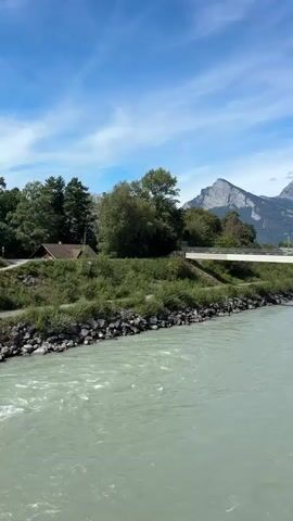 A vertical video shows a wide, fast-flowing river with greenish-blue water. The riverbanks are lined with rocks and green vegetation. In the background, a white bridge spans the river, supported by pillars. Beyond the bridge, a treeline separates the river from a mountain range with distinct peaks, one of which is a prominent jagged rock formation. The sky above is blue with scattered white clouds. A small, dark-colored building with a brown roof is visible among the trees on the left bank. There are no people or animals visible in the video. The overall scene depicts a natural landscape with a man-made structure, likely a rural or mountainous region.