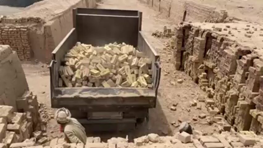 A truck bed full of bricks is being unloaded by two men into an area surrounded by crumbling brick walls. One man is wearing a white turban and beige clothes, and the other has a cap and light shirt. Bricks are scattered all around the ground. It appears to be a hot, sunny day. The surrounding area looks like an archaeological site or abandoned brickyard.