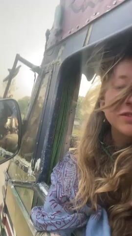 A young woman with long, wavy blonde hair is filming herself from inside a moving vehicle. She is wearing a light blue patterned shirt and has tattoos on her hands and arm. The vehicle appears to be an older, rugged type, possibly a truck, and she is looking out an open window with her hair blowing in the wind. Other people are visible in the interior of the vehicle. She smiles briefly. The background outside the window shows a road and landscape.