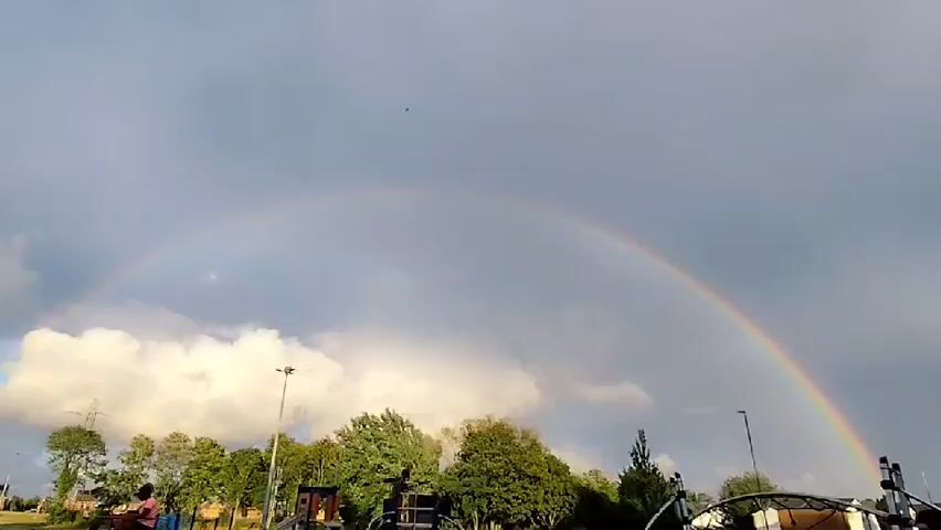 The video opens with a wide shot of a cloudy sky with a prominent rainbow arcing across it. In the foreground, silhouetted figures of people and parts of a structure are visible. As the video progresses, the camera zooms in on the sky, focusing on a small airplane flying higher up. The airplane is a dark silhouette against the gray sky. The rainbow remains visible in the background throughout the clip. The environment appears to be outdoors, possibly a park or open area, with trees and utility poles visible at the bottom of the frame in the initial shot.