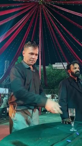A video depicts two men standing behind a table. The man on the left is fair-skinned with short dark hair and is wearing sunglasses on top of his head. He is wearing a dark long-sleeved shirt and light-colored pants. He is manipulating a deck of cards in his hands. To his right, a man with darker skin and a beard is wearing a black suit jacket. He is resting his hand on the table, near a glass of red liquid. The table is covered in a green cloth. The background shows a partially visible outdoor setting with trees, other people, vehicles, and buildings in the distance. Above them is a decorative covering of red and white draped fabric with strings of lights.