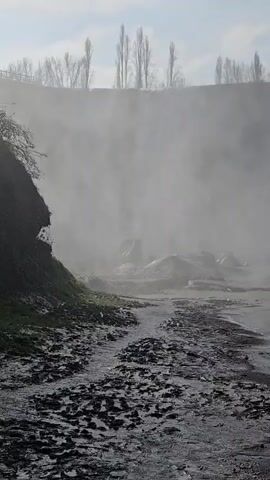The video shows a waterfall obscured by mist. The bottom of the falls is visible, with water flowing over rocks and onto a dark, muddy shore. A turtle swims in the water. A line of trees is silhouetted against the sky in the background. The sky is blue with some clouds.