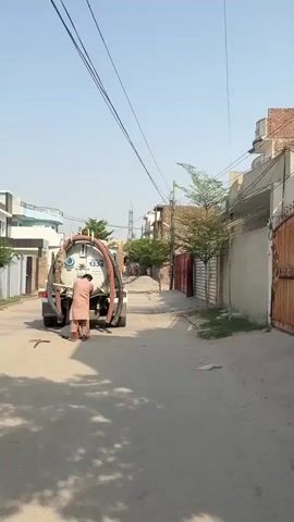 A man wearing a turban and peach-colored shalwar kameez stands near a sewage truck on a paved street and places a corrugated hose into a sewer hole. A crowbar lies next to the sewer hole. The white truck has blue lettering and features portraits on the front. The video pans from the rear of the truck to the tank, and then to the front of the vehicle. Houses border both sides of the street. The sky is clear and bright.