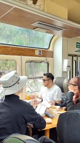 Three people are sitting at a wooden table on a train. They are eating from containers. A man wearing a white t-shirt and sunglasses is looking out the window. A woman with red hair in a ponytail and sunglasses is resting her chin on her hand. A person wearing a hat and a black shirt is looking at their phone. The train is moving, and trees can be seen outside the window. An employee in an orange vest walks down the aisle. Other passengers are visible further down the train. There is a sign that says "Salida Exit" above a door.