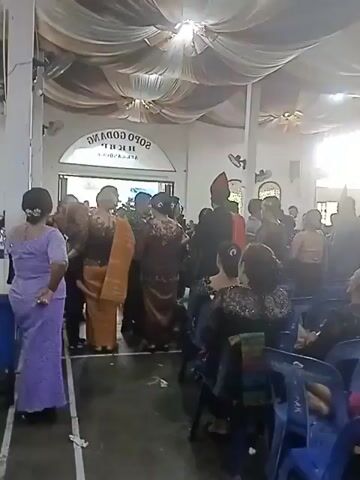 A video shows an indoor gathering of people, possibly a wedding or religious ceremony. The room is decorated with draped fabric on the ceiling. In the background, a sign is visible with text that reads "SOPO GODANG HKBP". Several individuals are dressed in traditional attire, including women in long dresses and men in suits or formal wear. Some people are seated, while others are standing and conversing. The overall atmosphere appears to be festive or solemn, depending on the context of the event.