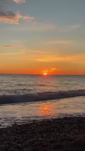 The video shows a sunset over a body of water. The sun is setting on the horizon, casting an orange glow over the water and sky. Small waves are lapping against a pebble beach. The sky has patches of blue and pink. There are no people visible in the video.