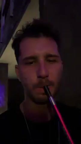 A close-up shot shows a man with dark hair and a mustache smoking a hookah. He inhales from the hookah hose and exhales a cloud of smoke. The scene is dimly lit with purple hues. He is wearing a black t-shirt and a silver chain. The background is blurred. The hookah hose has a red glow.